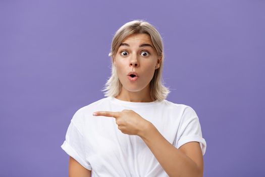 Waist-up shot of overwhelmed excited and surprised attractive urban female in white t-shirt folding lips gasping from amazement being questioned asking question and pointing left over purple wall. Copy space