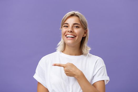 Close-up shot of amused happy and entertained good-looking sociable woman with fair hair and beautiful smile grinning while pointing left with index finger showing awesome copy space over purple wall. Emotions concept