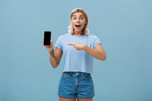 Studio shot of amazed and impressed charming european blonde in casual clothes gasping charmed at camera while showing smartphone screen pointing at device with finger over blue background. Technology and advertisement concept