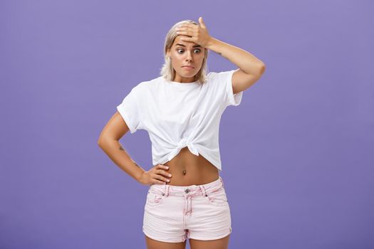 Woman got in serious bad sitation feeling concerned and nervous starting panic holding hand on forehead pursing lips staring down with shocked, perplexed expression being in trouble over purple wall.