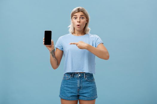 Wow look what found online. Portrait of impressed speechless good-looking young sportswoman in t-shirt and denim shorts saying wow folding lips showing smartphone and pointing at screen over blue wall. Technology concept