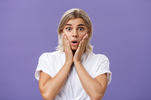 Waist-up shot of stunned impressed gorgeous tanned female in white t-shirt with blond hair gasping with opened mouth from surprise holding hands on cheeks from amazement and curiosity. Emotions concept