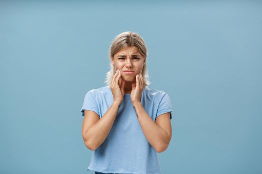 Girl feeling pain in teeth after drinking hot coffee. Displeased unhappy troubled young girl with blond hair frowning feeling discomfort in mouth holding hands on cheeks suffering toothache.