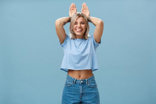 Time to relax. Portrait of happy playful cute blonde girl in trendy t-shirt with pierced belly and tattoo smiling joyfully holding palms on head like bunny ears having fun over blue background. Emotions concept