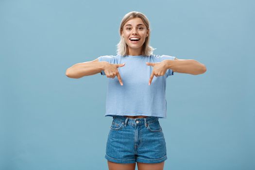 You have to see this. Portrait of joyful amused and happy stylish blonde female in trendy t-shirt pointing down with arms near chest smiling broadly showing amazing copy space over blue background.