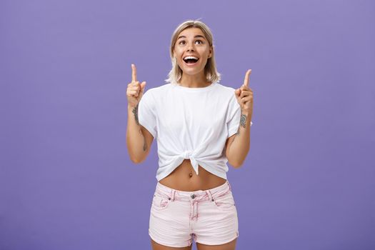 Lifestyle. Woman seeing miracle being impressed and delighted like child staring and pointing up with broad thrilled smile standing over purple background in summer trendy outfit being joyful and excited.