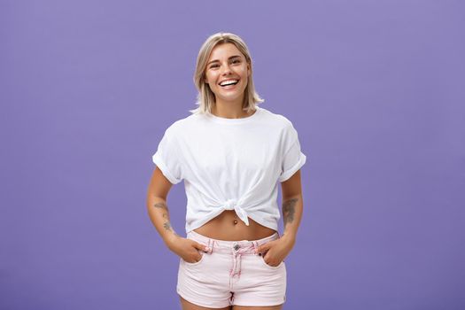 Lifestyle. Joyful attractive tanned girlfriend with blond hair and tattoos on arms with cropped top chilling by pool with friends talking with amused happy expression smiling at camera holding hands in pockets.