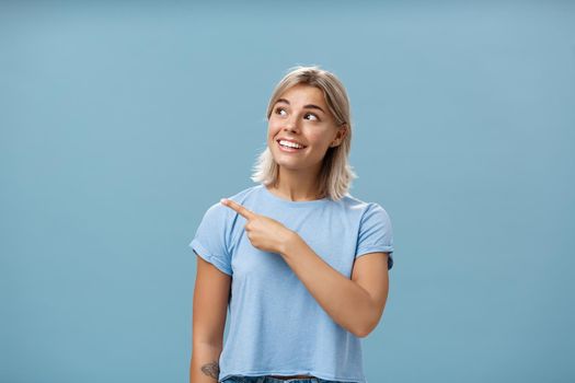 Lifestyle. Waist-up shot of creative and dreamy happy attractive blonde female in casual t-shirt looking and pointing at upper left corner with admiration and joy standing over blue background amused and curious.
