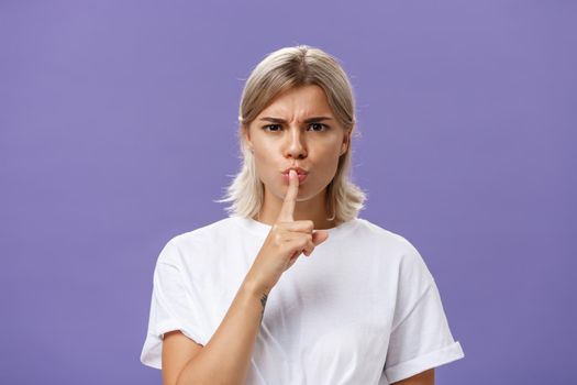 Lifestyle. Close-up shot of serious strict good-looking caucasian female with blond hair shushing at camera displeased and disappointed frowning holding index finger over folded lips in shush sign.