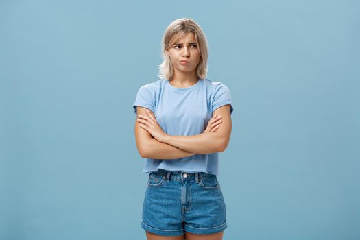 Uncertain troubled and perplexed attractive blond woman with tanned skin holding hands crossed on chest pouting and frowning looking right with worried unsure and doubtful look over blue wall. Feelings and emotions concept