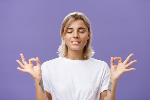 Peaceful calm good-looking female blonde in white t-shirt closing eyes smiling relieved and happy feeling satisfied with life standing in lotus pose with zen gesture over purple background. Copy space
