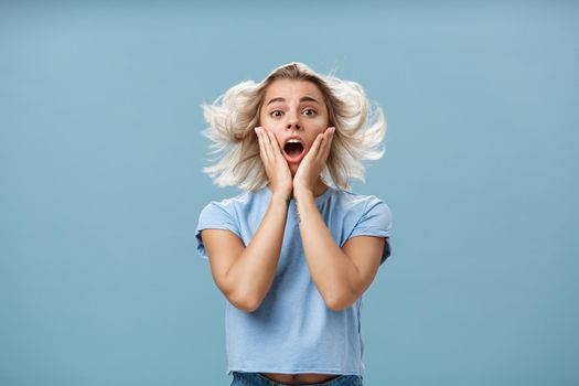 Indoor shot of shocked and impressed young stylish european female with blond hair holding hands on cheek from empathy and surprise opening mouth facing wind posing with hairstyle floating in air. Copy space