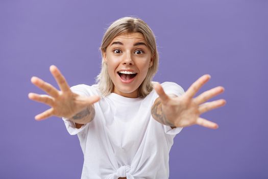 Lifestyle. Waist-up shot of amused and excited attractive stylish young woman in white t-shirt pulling hands at camera with desire smiling thrilled and happy wanting hug or take something over purple background.