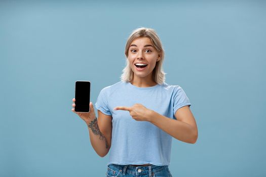 Lifestyle. Waist-up shot of thrilled and impressed good-looking female student in casual t-shirt smiling joyfully pointing at smartphone screen showing awesome place via internet to friend over blue background.