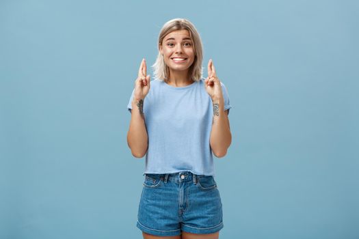 Hope dream come true soon. Portrait of emotive happy and optimistic attractive young european woman with blond hair and tanned skin crossing fingers for good luck and smiling over blue wall.