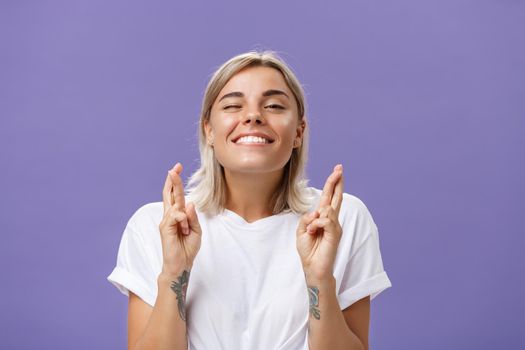 Waist-up shot of hopeful optimistic attractive stylish woman in white t-shirt with tattooed arms winking smiling joyfully while crossing fingers for good luck making wish over purple background. Body language concept