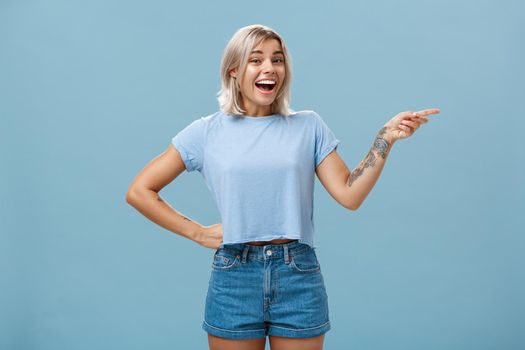 Studio shot of impressed and surprised entertained blonde female in summer outfit holding hand on waist pointing right and smiling joyfully being amazed and amused over blue background. Emotions and advertisement concept