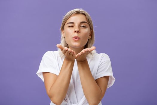 Blowing sweet kiss to all fans. Flirty tender and cute stylish european female with blond hair and tanned skin holding palms near mouth folding lips while sending mwah at camera over purple wall.