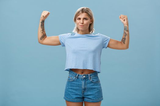 Lifestyle. Strong and powerful good-looking sportswomen with tattoos in t-shirt and shorts raising arms showing big muscles and biceps smiling proudly while bragging with physical strength over blue wall.