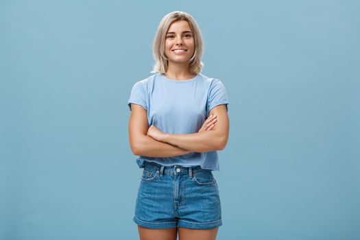Ambitious good-looking young tanned female with blond hair standing in confident pose with hands crossed on chest smiling broadly standing in denim shorts over blue background. Copy space