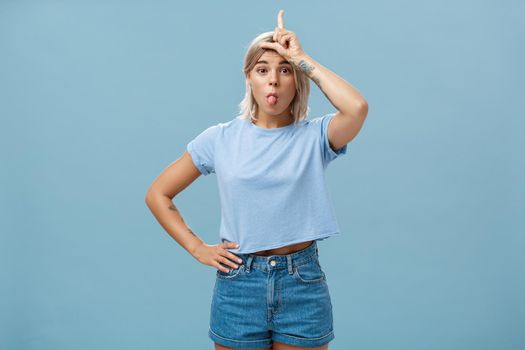 Lifestyle. Studio shot of playful triumphing good-looking blonde female with tattoed arms showing letter L on forehead and sticking out tongue mocking over loser and celebrating own victory over blue wall.