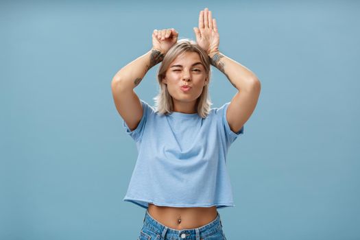 Lifestyle. Flirty attractive and stylish cool girlfriend with tattooed arms and pierced belly winking folding lips in air kiss showing bunny ears with palms on head standing over blue background playful.