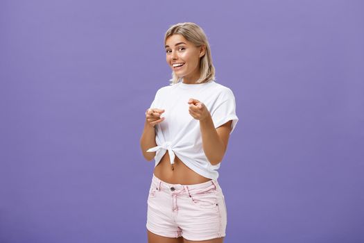 We need you in team. Joyful friendly and good-looking blonde female student in white t-shirt and shorts pointing with finger guns at camera and smiling broadly greeting friend over purple wall. Copy space