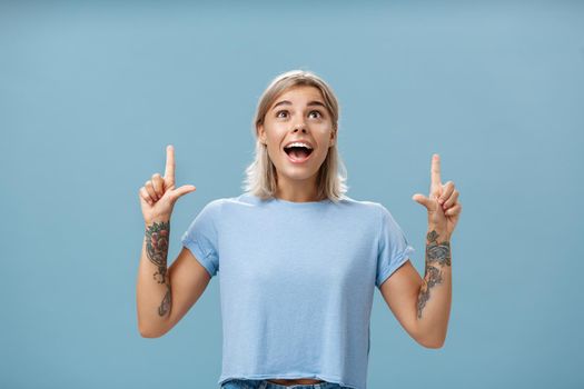 Studio shot of impressed speechless happy good-looking woman with tattoos on arms dropping jaw from amazement and joy gazing fascinated and pointing up standing over blue background. Emotions concept