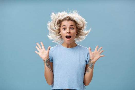 Waist-up shot of impressed surprised attractive and cute girl with blonde hair floating in air and tattooed arm jumping with spread raised palms and opened mouth over blue background. Emotions concept