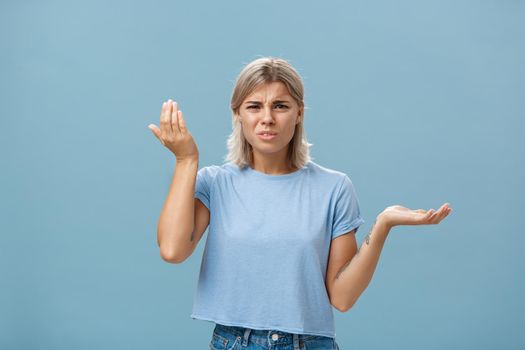 Wtf you saying. Portrait of pissed and confused stylish girlfriend with blond hair raising palm high and aside with perplexed expression being questioned hearing stupid nonsense over blue wall.