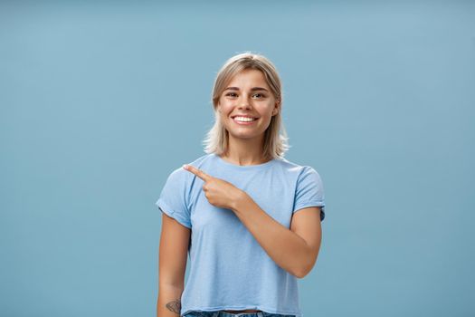 Come with me. Portrait of stylish pleased good-looking female student with blond hair in trendy t-shirt pointing at upper left corner and smiling with delight and happiness posing over blue background.