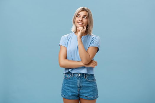 Dreamy thoughtful and creative artistic blonde woman in denim shorts and t-shirt smiling curiously touching lip and gazing at upper left corner feeling nostalgic, thinking over blue wall.