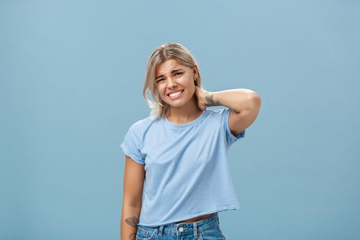 Lifestyle. Girl unwilling to go with friend feeling awkward and unsure how say no frowning and clenching teeth making apologizing face rubbing neck behind saying sorry while rejecting offer over blue background.