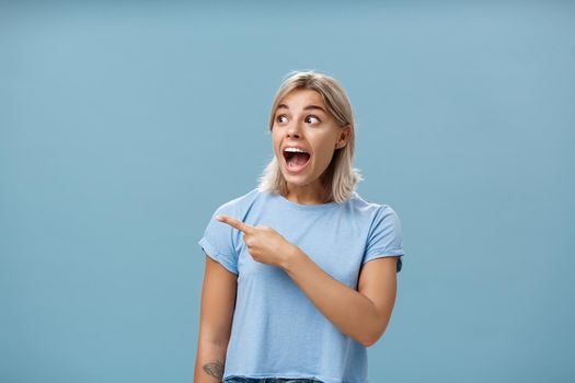 Girl being charmed and excited visiting awesome event pointing and staring left amazed and delighted standing happy and overwhelmed over blue background in trendy outfit dropping jaw. Emotions and advertising concept