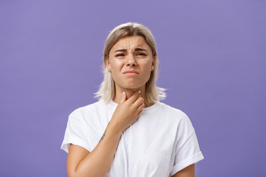 Displeased young stylish caucasian woman with blond hair and tanned skin feeling discomfort in throat having inflammation or feeling cold touching neck and frowning dissatisfied from pain. Copy space