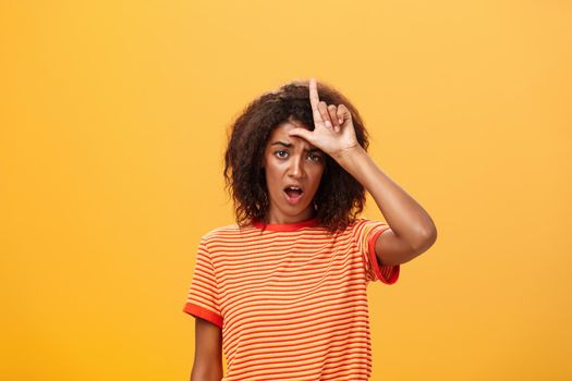 Girl thinks she loser. Portrait of gloomy bothered and displeased african american woman with afro hairstyle showing l word over forehead complaining feeling gloomy and unhappy over orange background.