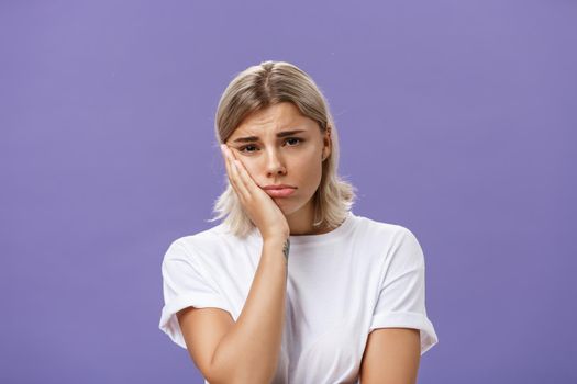 Waist-up shot of unhappy miserable and sad cute blonde female in white casual t-shirt pursing lips leaning face on palm and frowning from disappointement and regret over purple background. Copy space