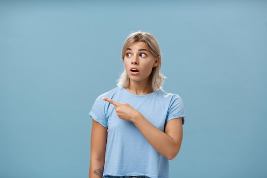 Waist-up shot of speechless shocked and impressed attractive young woman with blond hair dropping jaw staring and pointing confused and shook left standing over blue background. Emotions concept