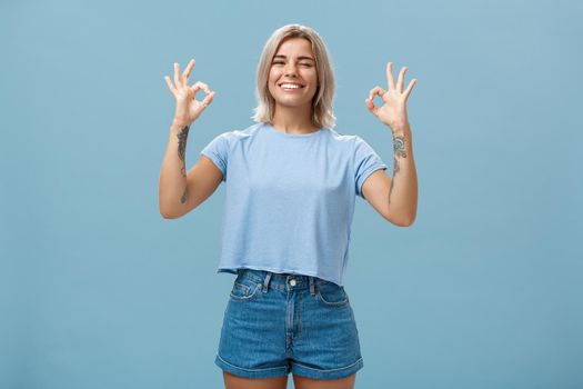 I got you covered. Enthusiastic attractive and energized female blonde with tattooed arms showing okay or perfect sign and winking to assure friend everything done smiling joyfully over blue wall.