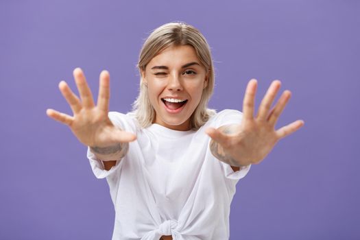 Reaching to you. Portrait of flirty good-looking and confident stylish woman with tattoos on arms winking sticking out tongue playfully and smiling pulling hands towards camera over purple background. Emotions and body language concept