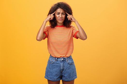 Girl cannot concentrate and think straight being loaded with bad thoughts looking concerned and intense, frowning holding fingers on temples suffering disorientation and migraine over orange wall. Emotions and health concept