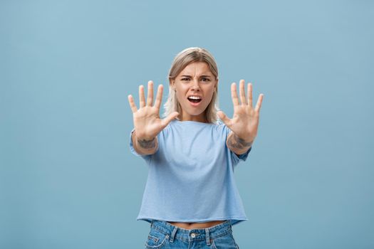 Hold right there. Portrait of intense displeased and irritated attractive young female in blue t-shirt pulling hands towards camera in stop or not gesture frowning and making annoyed expression.