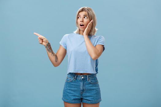 Studio shot of nervous shocked good-looking young woman with fair hair and tatooed arm gasping pointing and looking left with worry holding hand on cheek posing over blue background. Emotions and advertisement concept