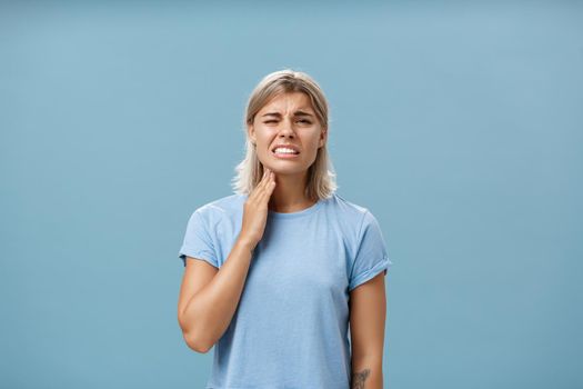 Ouch my throat. Displeased unhappy good-looking adult blonde girl with short haircut clenching teeth from painful feeling touching neck having angina or inflammation posing over blue background. Health concept