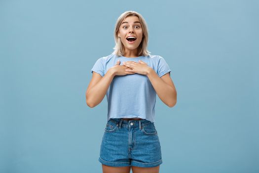 Lifestyle. Portrait of amazed and charmed attractive blond female student with tanned skin in denim shorts and summer t-shirt holding palms on breast gasping and smiling joyfully being grateful and pleased.