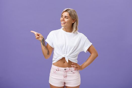 Hey nice job. Pleased carefree good-looking and confident female shop manager in stylish outfit holding hand on waist pointing and looking left with satisfied and delighted look over purple wall.