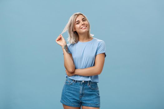 Studio shot of relaxed and chill popular attractive woman with blond hair and tattoos playing with strand tilting head and smiling as if listening boyfriend or flirting standing amused over blue wall. Body language and emotions concept