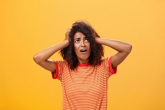 Displeased moody african girlfriend holding hands on head and frowning looking concerned and dissatisfied up in sky panicking rain will spoil hairstyle standing nervous over orange background. Lifestyle.