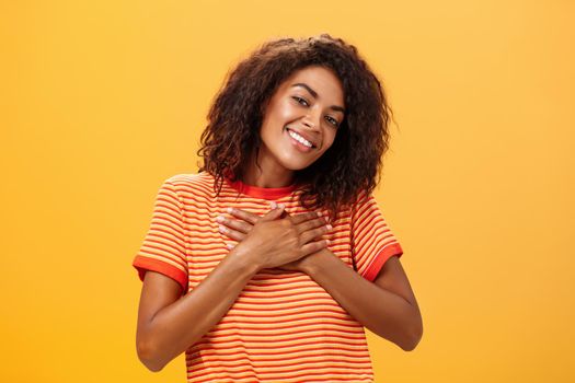 Waist-up shot of charming sensual dark-skinned woman with curly hairstyle in trendy t-shirt holding palms on heart looking with pleased loving smile feeling grateful and delighted over orange wall. Lifestyle.