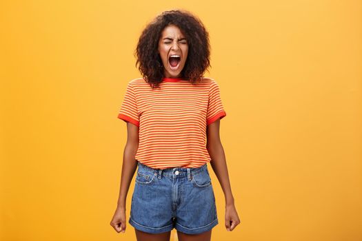 Girl feeling distressed and tensed losing temper and control of feelings yelling out loud clenching fists closing eyes standing intense and depressed over orange background screaming from rage. Emotions concept